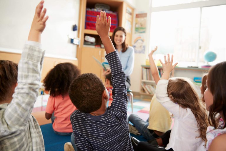 An image of seated pre-k students raising their hands.