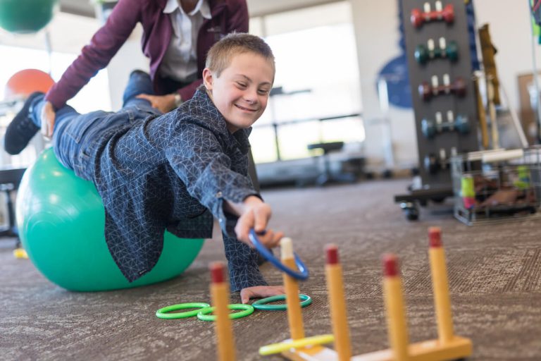 A patient attempting to place a grab a ring off of a peg while having their feet held over an exercise ball.