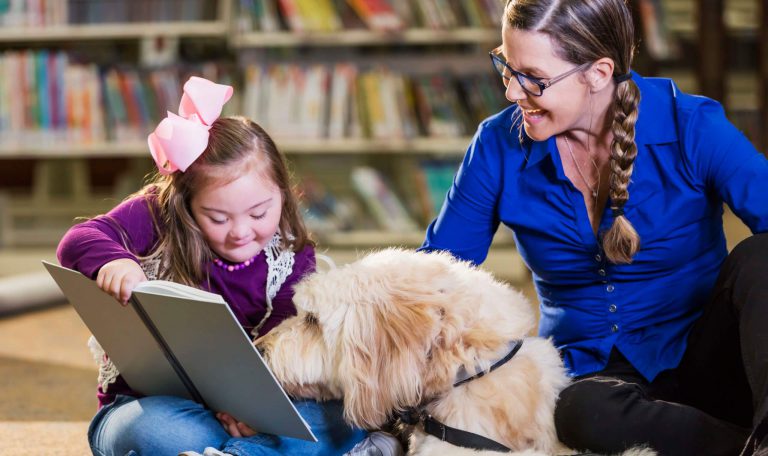 An image of a girl reading a book to a therapy dog in a library.