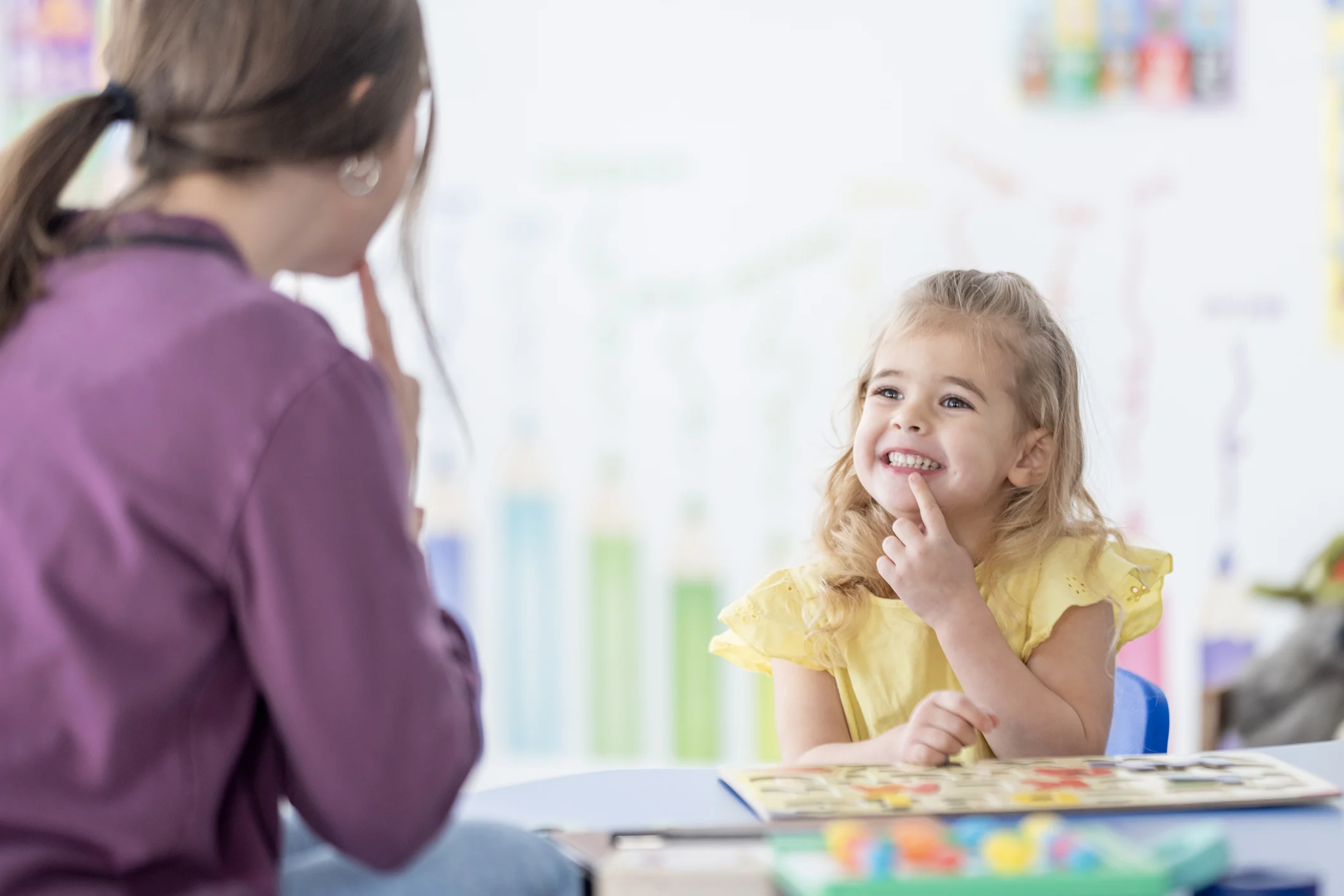 A smiling young girl in a yellow dress sits at a table with a colorful book while an adult woman teacher sits nearby in a purple top, fostering learning and playful conversation in a classroom.