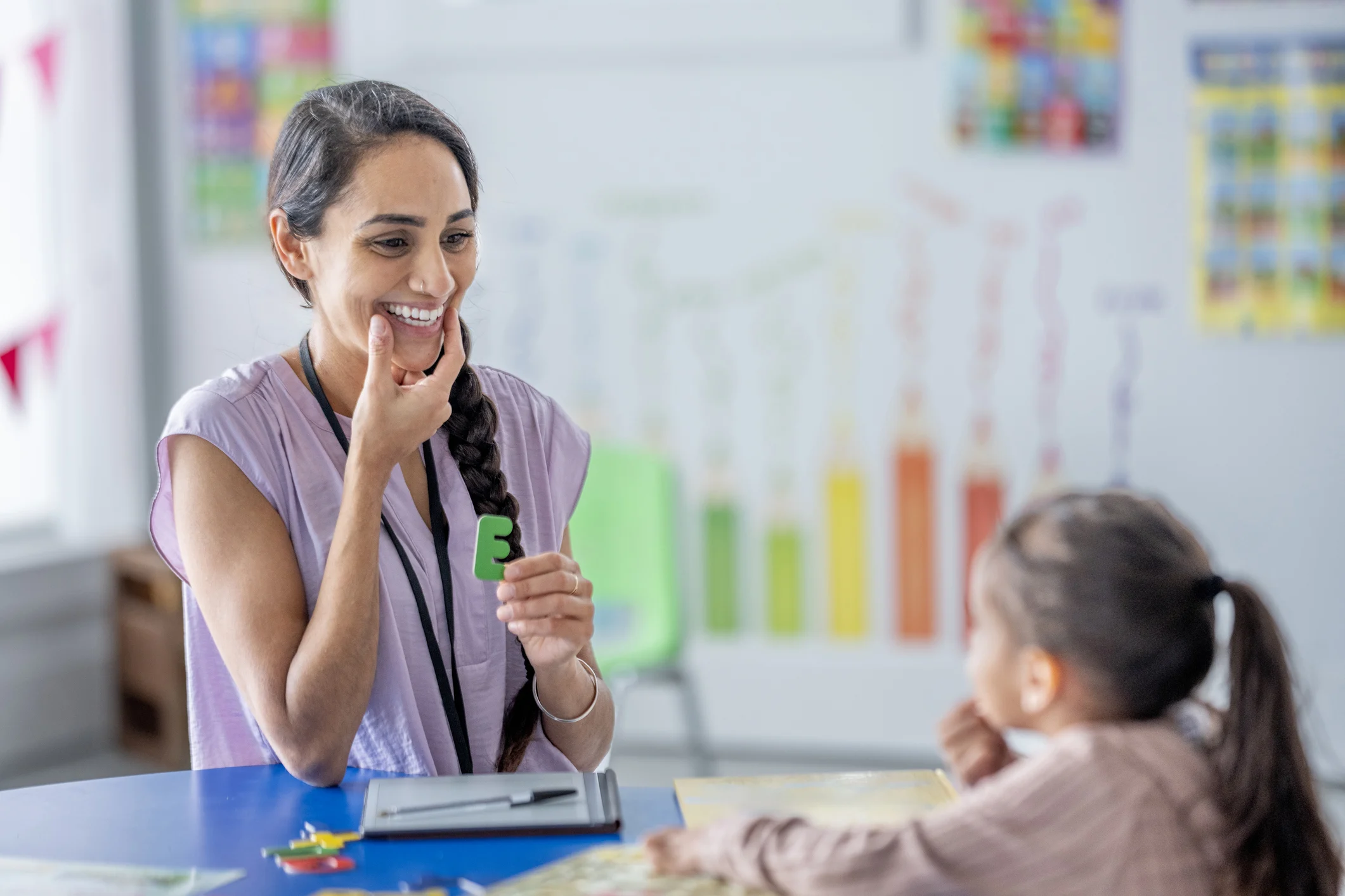 A Hispanic teacher smiles warmly as she engages a young student at a table filled with learning tools in a bright classroom, surrounded by colorful charts and educational decor that foster a positive learning environment.