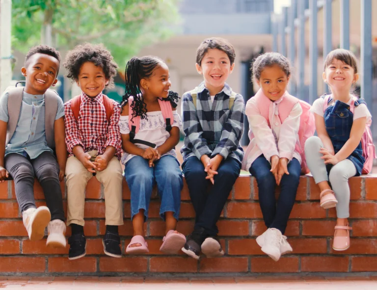 Portrait Of Smiling Elementary School Kids Sitting On Wall Outdoors At School